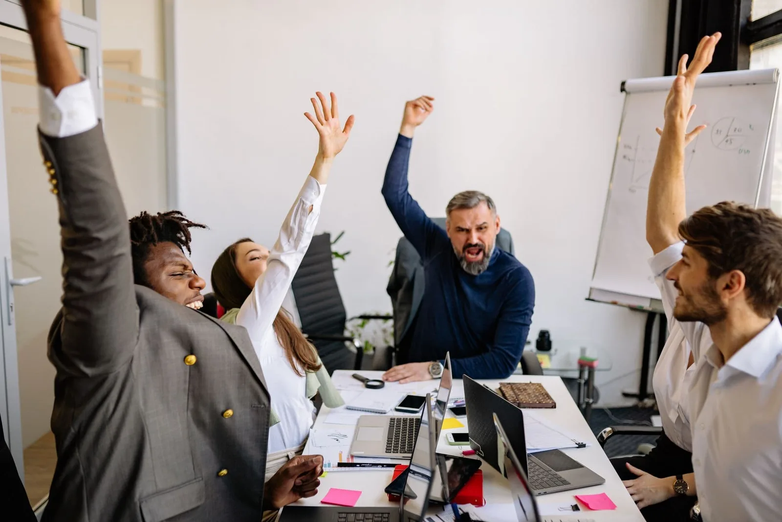 A team cheers with raised arms around a meeting table with laptops, sticky notes and a flip chart